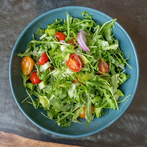 A vibrant arugula salad with cherry tomatoes, cucumber, and red onion in a teal bowl.