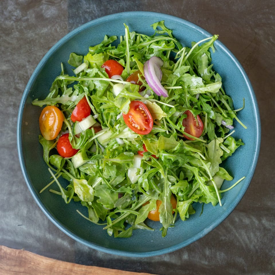 A vibrant arugula salad with cherry tomatoes, cucumber, and red onion in a teal bowl.