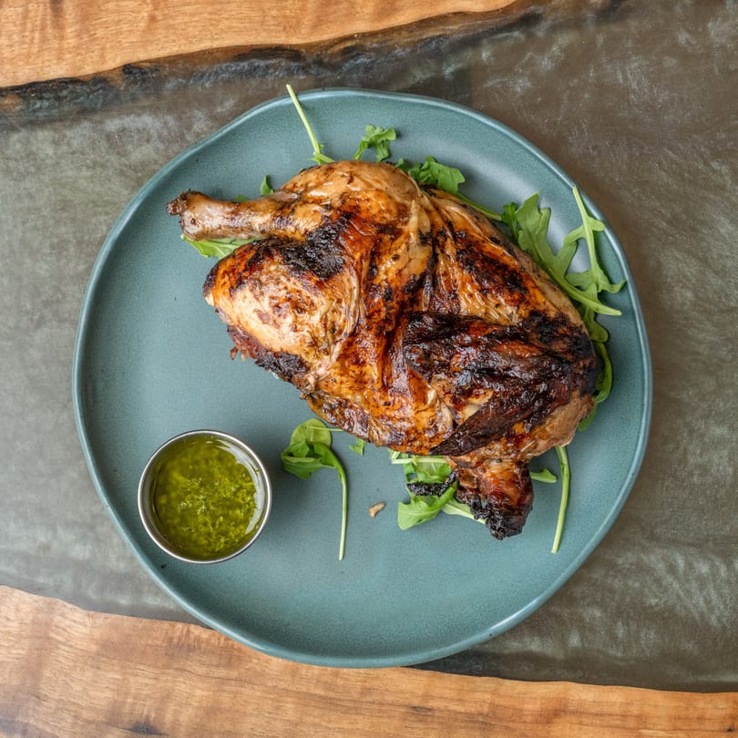 Grilled chicken with crispy skin, served on a bed of arugula with a side of green sauce.