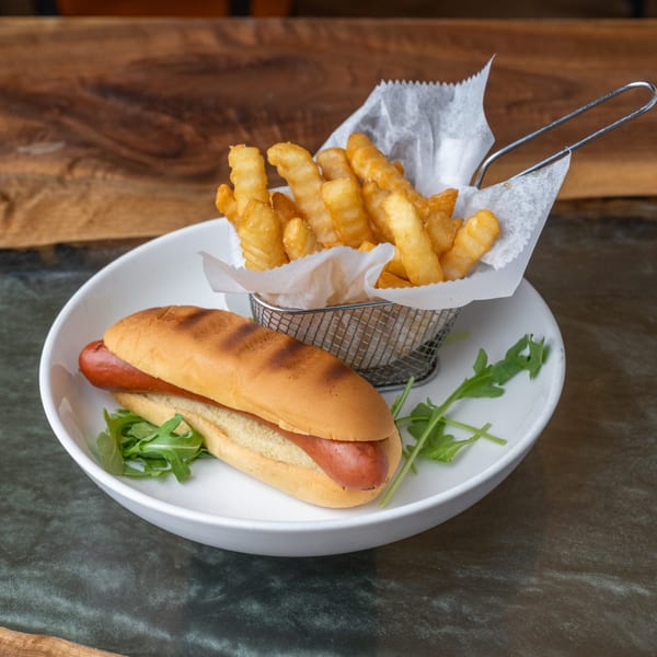 A grilled hot dog in a bun with arugula, served with crinkle-cut fries in a metal basket.