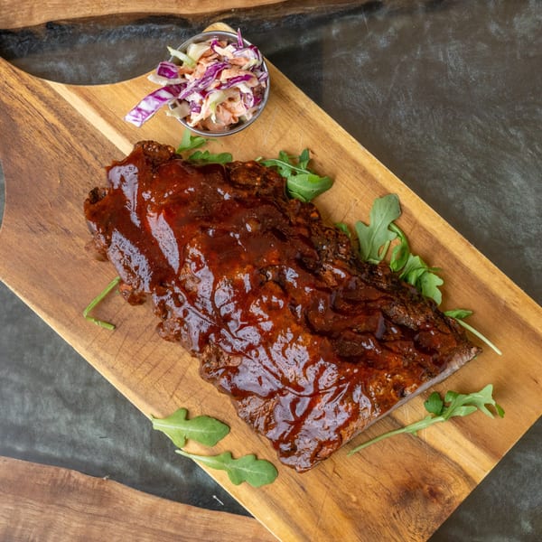 Glazed BBQ ribs with a side of coleslaw and arugula on a wooden cutting board.