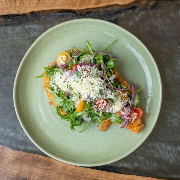 Crispy chicken schnitzel topped with arugula, cherry tomatoes, red onion, and shredded cheese on a green plate.