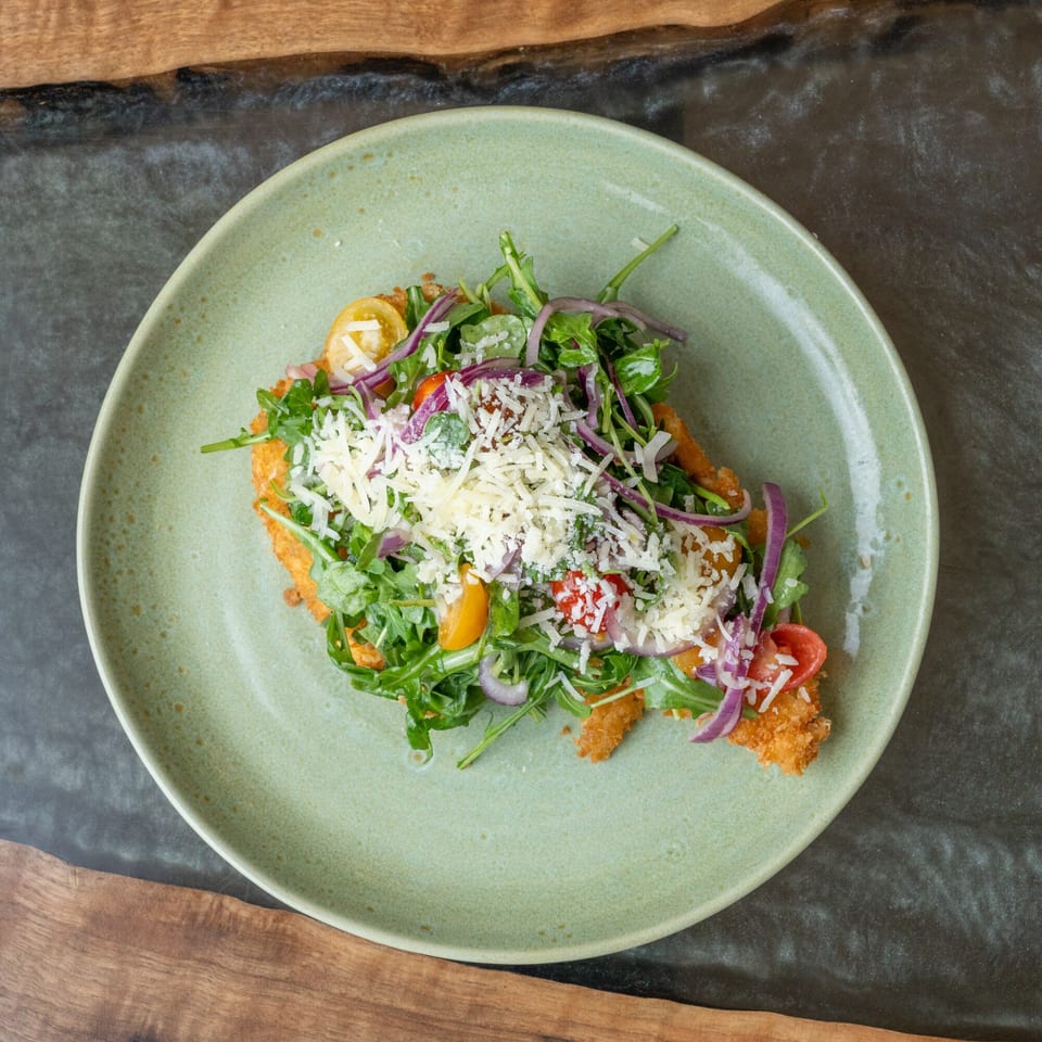 Crispy chicken schnitzel topped with arugula, cherry tomatoes, red onion, and shredded cheese on a green plate.