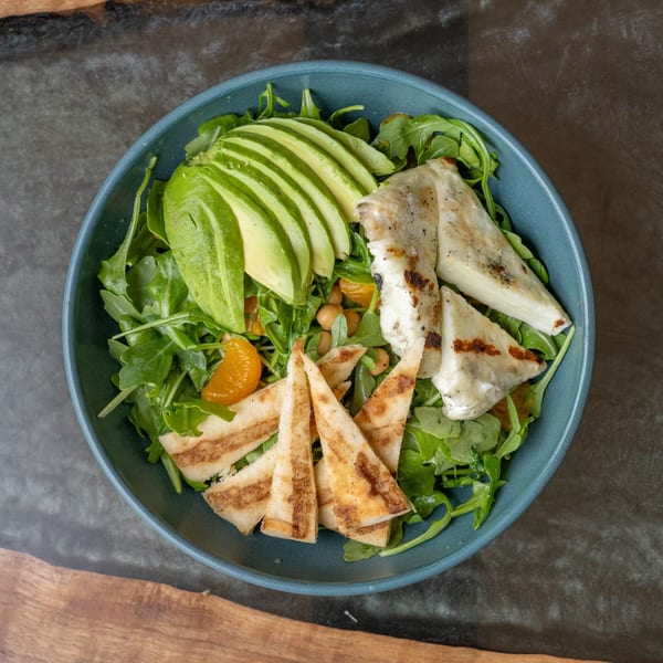 Overhead view of a vibrant salad with sliced avocado, grilled chicken, and pita bread in a blue bowl.