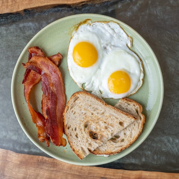A light green plate holds two fried eggs, crispy bacon strips, and two slices of toast.