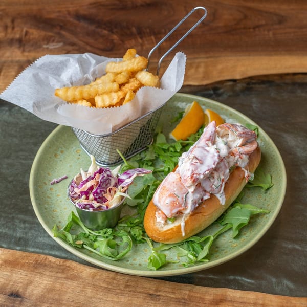 Lobster roll with creamy dressing, crinkle-cut fries, coleslaw, and orange slices on a green plate.