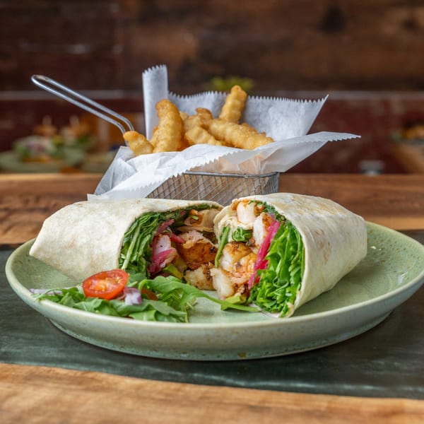 A shrimp wrap, cut in half, with a side of fries in a metal basket on a wooden table.