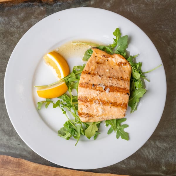 Grilled salmon fillet with lemon wedges and arugula on a white plate.